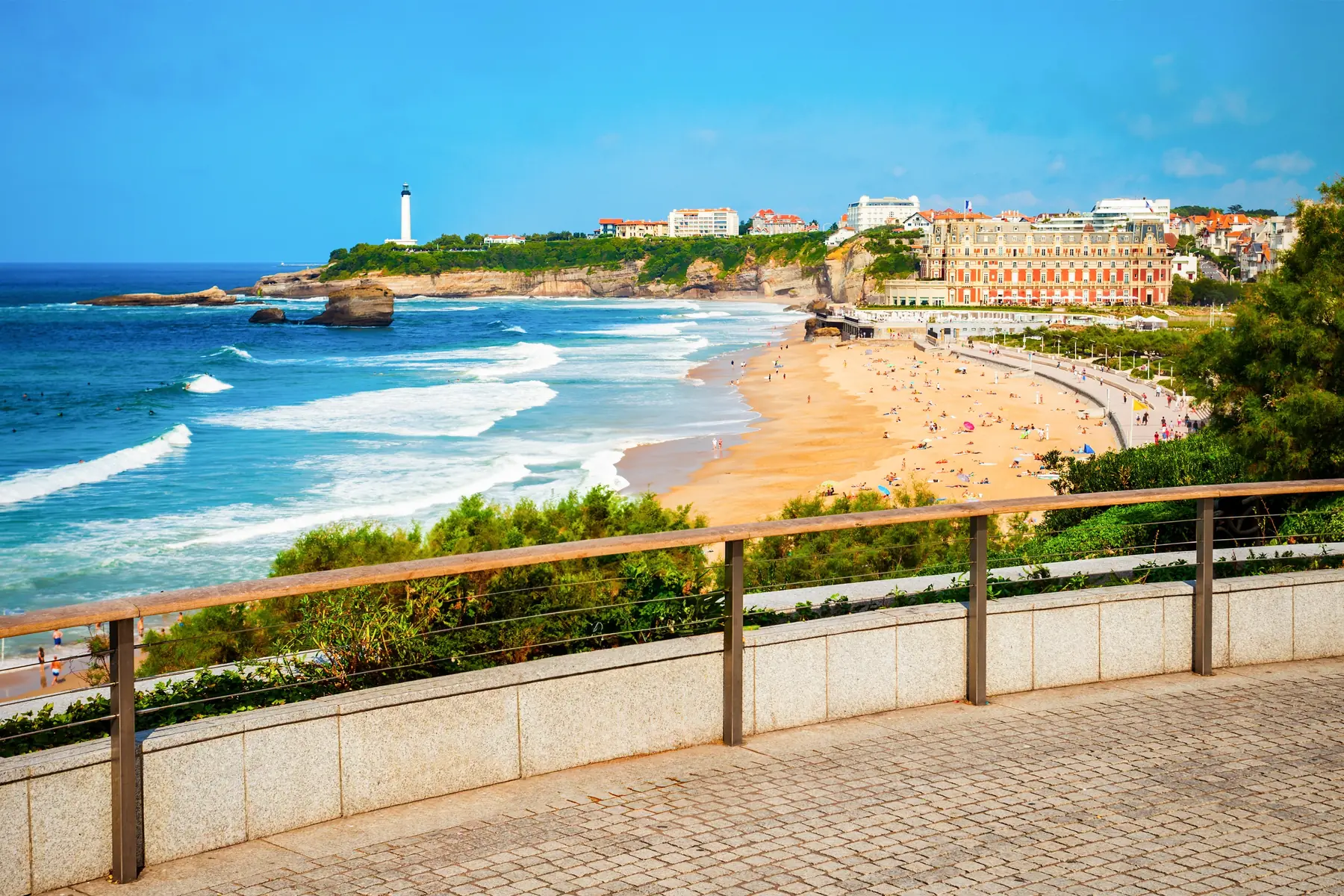 View from a promenade over a wide sandy beach, the ocean and a seaside town, with a white lighthouse on the headland