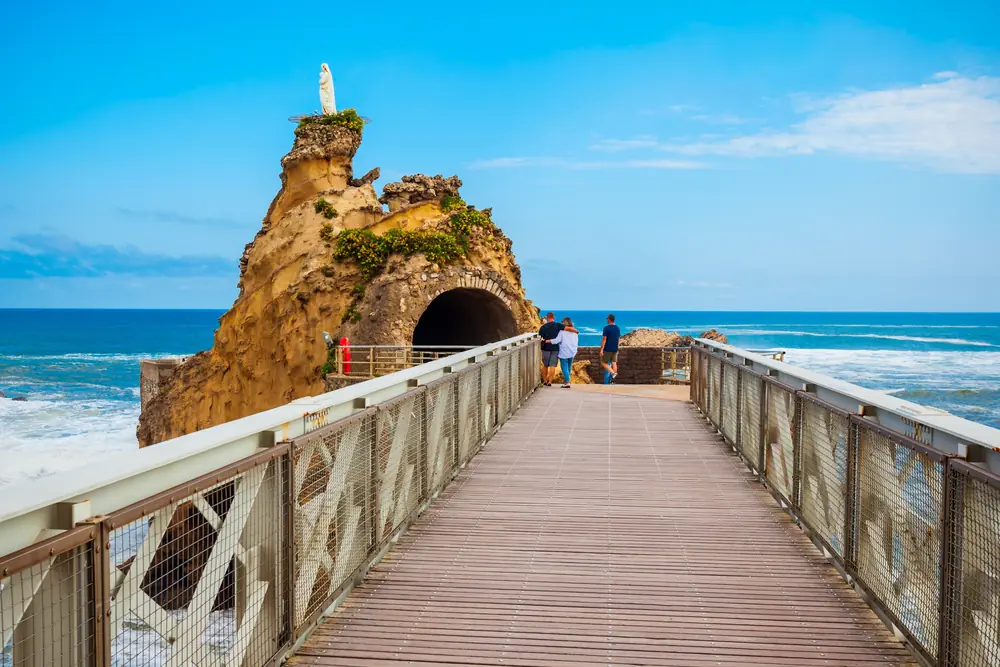 A wooden and metal footbridge leading to a rock topped by a statue above the ocean