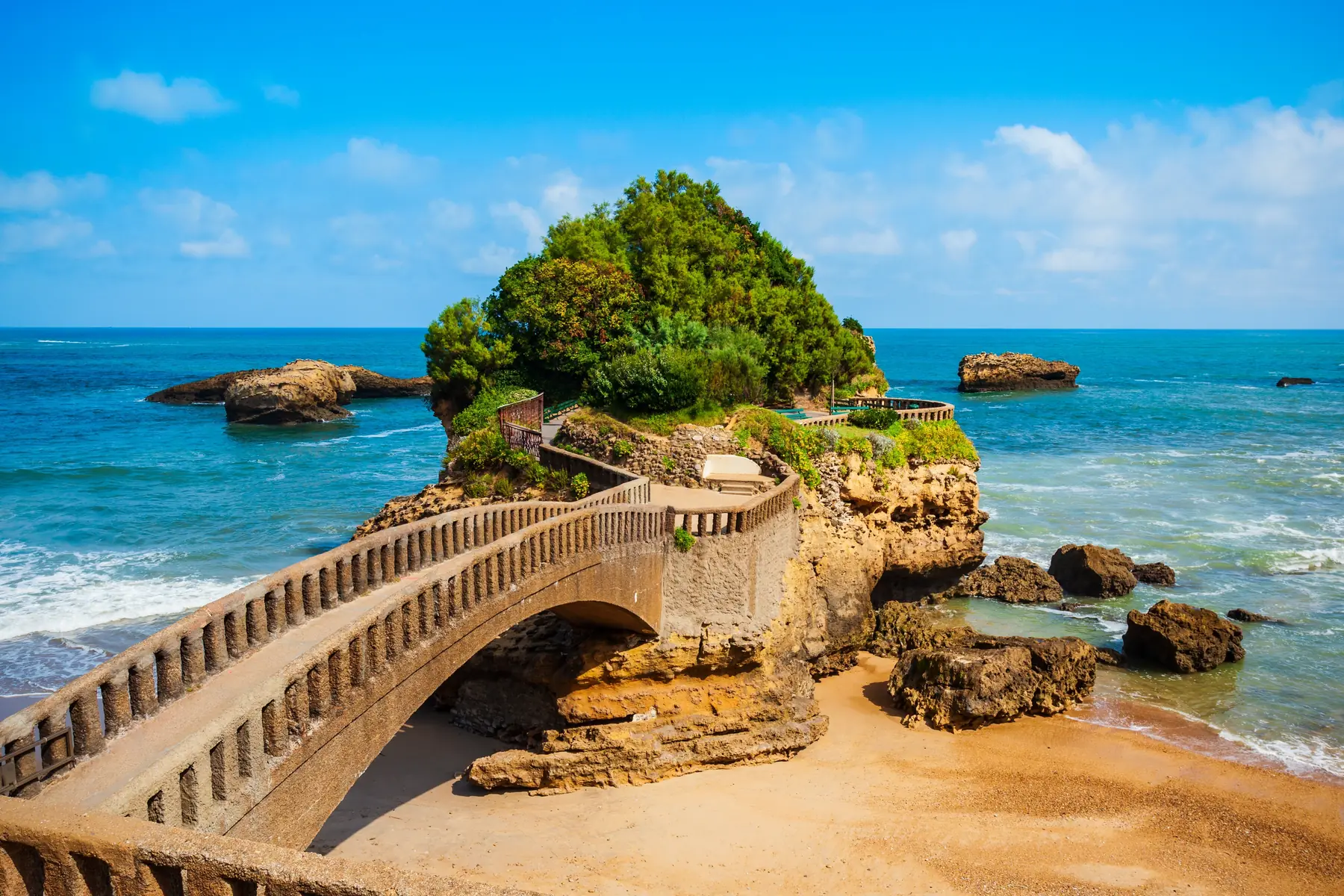 A stone bridge connects the beach to a tree-covered rock above turquoise water